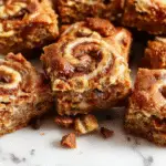 A close-up of a freshly baked cinnamon swirl snickerdoodle blondie bar, showcasing its golden-brown top with cinnamon swirls and a slightly crackled surface. The edges are slightly crisp, with a soft, chewy center visible at the cut edge. The blondie is placed on a rustic wooden surface, garnished with a sprinkle of cinnamon and a few additional swirls visible within the cut piece.