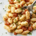 A creamy Tuscan butter beans dish served in a rustic white bowl, garnished with fresh herbs. The beans are coated in a rich, velvety sauce with glimpses of spinach and crispy bacon bits, surrounded by vibrant green beans on a wooden table with natural lighting.