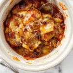 A warm, golden-brown caramel apple dump cake served in a rustic ceramic dish. Topped with a light drizzle of caramel sauce and garnished with chopped nuts and fresh apple slices. The dessert layers reveal tender apple pieces beneath a crumbly, cinnamon-spiced cake crust, all presented on a wooden table with autumn-themed decorations in the background.