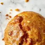 A golden-brown loaf of pumpkin sourdough bread with a rustic, slightly cracked crust, sliced to reveal its moist, orange-tinted crumb speckled with bits of pumpkin. The bread rests on a wooden cutting board, garnished with a few small pumpkin pieces and a sprig of fresh herbs on the side. The background features a cozy kitchen scene with warm lighting that highlights the bread's inviting texture and vibrant color.