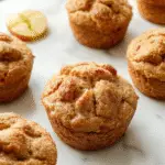 A close-up of a golden-brown apple cinnamon Greek yogurt muffin with a slightly cracked top, sprinkled with cinnamon and small apple pieces visible. The muffin is on a rustic wooden platter alongside fresh green apple slices and a cinnamon stick, with a blurred background of a cozy kitchen setting.