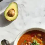 A steaming bowl of hearty meatless taco soup garnished with fresh cilantro, shredded cheese, and a lime wedge. The vibrant orange and red hues of beans, corn, and tomatoes contrast against the dark bowl, with a rustic wooden surface and scattered ingredients in the background.