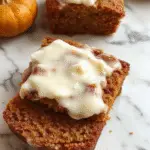 A sliced pumpkin bread loaf topped with rich, creamy cream cheese frosting, displaying a moist, dense texture with a golden-brown crust. The frosting is generously spread, with some parts slightly melting into the bread's surface, and the bread is presented on a rustic wooden platter with a few pumpkin seeds scattered around, highlighting the seasonal theme.