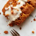 A loaf of pumpkin streusel bread resting on a rustic wooden surface, topped with a crunchy streusel crumb, with a slice of the bread revealing a moist, orange-centered crumb. The background features a warm, inviting setting with cinnamon sticks and fall leaves, emphasizing the cozy, seasonal vibe.