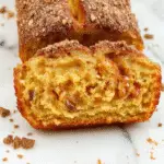 A golden-brown loaf cake topped with a shimmering cinnamon sugar crust, sliced to reveal a moist interior dotted with bits of spiced apples, garnished with a dusting of cinnamon sugar. The cake is presented on a rustic wooden board with a few apple slices and cinnamon sticks nearby, evoking warm fall flavors.