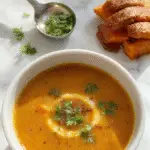 A vibrant bowl of Spicy Butternut Squash Sweet Potato Soup, garnished with a swirl of cream and a sprinkle of chili flakes. The soup has a smooth, velvety texture with a rich orange hue, served in a rustic ceramic bowl on a wooden table. Fresh herbs and roasted squash pieces are visible on top, with a spoon resting beside the bowl.