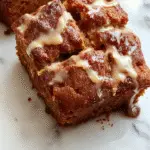 A freshly sliced pumpkin bread loaf with a golden-brown crust, topped with a drizzle of icing. The slices reveal moist, dense interior speckled with pumpkin seeds and spices, arranged on a rustic wooden surface alongside a cinnamon stick and a small bowl of creamy butter.