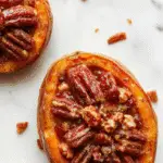 A close-up of a sliced twice-baked sweet potato topped with a caramelized pecan pecan crust, showcasing golden-brown edges, scattered chopped pecans, and a glossy glaze garnish. The dish is presented on a rustic wooden plate with a few whole pecans and a sprig of fresh herbs nearby, evoking warmth and comfort.