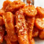 A close-up of a glossy, caramelized batch of candied carrots served on a rustic white plate. The carrots are vibrant orange, coated with a shiny brown sugar glaze, with some caramelized edges visible. The presentation is simple yet appetizing, highlighting the texture and richness of the candied carrots.