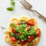 A bowl of vibrant spinach and ripe cherry tomato pasta topped with grated Parmesan cheese, basil leaves, and a drizzle of olive oil. The dish is arranged on a rustic wooden table, with a few halved cherry tomatoes and fresh spinach scattered around, highlighting the colorful ingredients and fresh presentation.