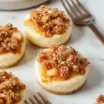 A close-up of mini cheesecakes topped with cinnamon-spiced apple slices and a caramel drizzle, arranged on a rustic wooden platter with a fall-themed background, showcasing golden-brown crusts and creamy filling amidst fresh apple slices and cinnamon sticks.