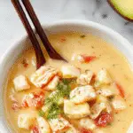 A bowl of creamy chicken noodle soup with tender chicken chunks, al dente noodles, and a rich, velvety broth garnished with chopped parsley, served on a rustic wooden table with a spoon resting beside the bowl.