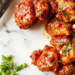 A crispy golden-brown General Tso's Chicken pieces piled on a white plate, garnished with chopped green scallions and a sprinkle of sesame seeds, with a glossy, tangy sauce drizzled over the chicken. The dish is presented with a side of steamed broccoli and white rice, creating a vibrant contrast of colors and textures, highlighting the crispy coating and saucy finish.