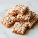 A close-up of golden-brown gingerbread-flavored Rice Krispie Treats shaped into squares, topped with a sprinkle of cinnamon. The treats are arranged on a festive red-and-green plaid paper, with some treat pieces stacked and slightly melting, showcasing their crispy texture with visible rice puff cereal.