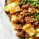 A vibrant stir-fry dish featuring tender ground beef and finely shredded cabbage, glazed with a savory sauce, served on a white plate with chopped green onions garnishing the top. The dish is arranged neatly, showcasing the colorful contrast between the beef's rich brown and the cabbage's pale green and white hues, with a hint of steam rising from the warm meal.