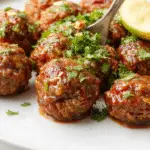 A close-up of a platter filled with glossy, grilled barbecue meatballs glazed with rich sauce, garnished with herbs, and arranged neatly on a rustic wooden board.