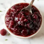 A glossy, vibrant red cranberry sauce served in a clear glass bowl topped with fresh rosemary sprigs, surrounded by whole cranberries and citrus slices on a rustic wooden table.
