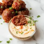 A close-up of a white plate featuring beautifully glazed Korean BBQ meatballs garnished with sesame seeds and chopped green onions. The meatballs are arranged neatly, showcasing their caramelized exterior and juicy interior. In the background, a small bowl of spicy mayo dip with a hint of red chili oil is visible, alongside a few fresh herbs for garnish.