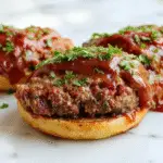 A close-up of golden-brown mini meatloaves baked in a muffin tin, each one perfectly shaped with a crispy top, garnished with a sprinkle of herbs. The mini meatloafs are arranged neatly on a white platter, with steam gently rising, showcasing their moist interior and textured crust. In the background, a rustic wooden table setting with fresh herbs and a side of vegetables adds warmth to the scene.