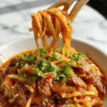 A close-up of a steaming bowl of Mongolian Ground Beef Noodles, featuring tender ground beef atop glossy, slightly sticky noodles, garnished with chopped green onions and sesame seeds, with a rich brown sauce coating the ingredients.