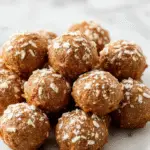 A close-up of a plate of golden-brown no bake pumpkin balls garnished with a sprinkle of cinnamon, arranged neatly on a white platter with a rustic fall-themed background, with some pumpkin puree and cinnamon sticks visible.