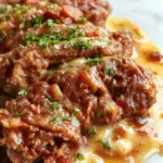A plated Salisbury steak dish featuring a perfectly seared beef patty topped with rich brown gravy, garnished with chopped parsley. The steak rests on a bed of creamy mashed potatoes, with a side of steamed green beans. The plate is white, accentuating the deep, savory colors of the dish, with a rustic wooden table background.