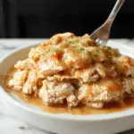 A close-up of shredded chicken coated in rich, glossy gravy served in a rustic white bowl. The shredded chicken is golden brown with tender textures visible, topped with fresh herbs. The background features a warm, inviting table setting with side dishes and a spoon resting beside the bowl.