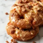 A sliced loaf of spiced apple cider donut bread resting on a rustic wooden board, with a golden-brown crust sprinkled with cinnamon sugar, surrounded by fresh apple slices and cinnamon sticks. The interior reveals moist, slow-rising crumb with visible apple chunks and a hint of spice, presented with a drizzle of caramel glaze on top.