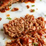 A perfectly baked beef meatloaf glazed with a rich, glossy sauce, sliced to reveal a moist, hearty interior with visible herbs and breadcrumbs, served on a rustic wooden platter with fresh herbs and roasted vegetables around it.