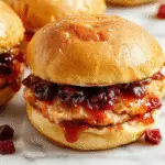 Close-up of golden-brown Hawaiian roll sliders filled with sliced turkey, vibrant red cranberry sauce, and melted havarti cheese. The sliders are garnished with poppy seeds and arranged on a rustic platter, with some cranberry sauce oozing slightly from the sides, showcasing a festive and appetizing presentation.