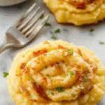 A creamy mound of vegan caramelized onion mashed potatoes topped with golden-brown caramelized onions and fresh herbs, served on a rustic ceramic plate with a sprig of rosemary and a drizzle of olive oil. The background features a cozy wooden table with fall-themed decorations and a side of green vegetables.